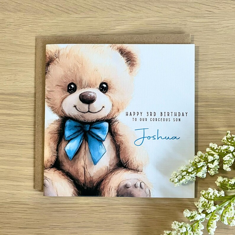 May include: A square greeting card featuring a tan teddy bear with a bright blue bow tie. The text reads Happy 3rd Birthday to our gorgeous son and the name Joshua in blue script. The card is set against a light background with a brown envelope.