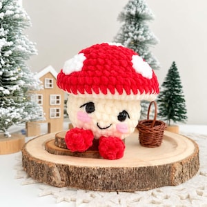 May include: A crocheted mushroom plush toy featuring a bright red cap with white spots, a cream-colored stem, black safety eyes, and pink embroidered cheeks. The plush has two small red feet and is accompanied by a miniature brown woven basket.