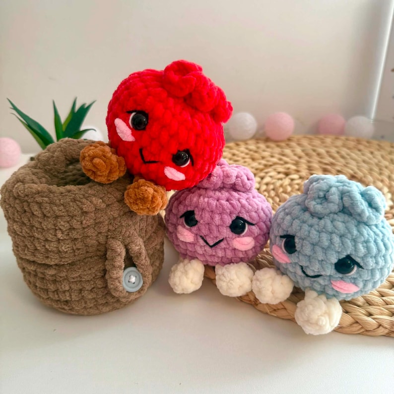 Puede incluir: Tres peque&ntilde;as figuras de peluche tejidas a ganchillo, redondas, con ojos de pl&aacute;stico negro y mejillas bordadas en rosa. Una es roja con pies marrones, una morada clara con pies blancos y una azul claro con pies blancos. Una cesta de ganchillo marr&oacute;n sostiene la figura roja.