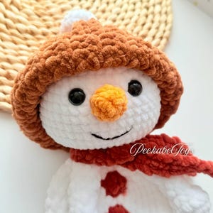 May include: A white crocheted snowman with a brown crocheted hat, a red crocheted scarf, and orange crocheted nose. The snowman has two red buttons.