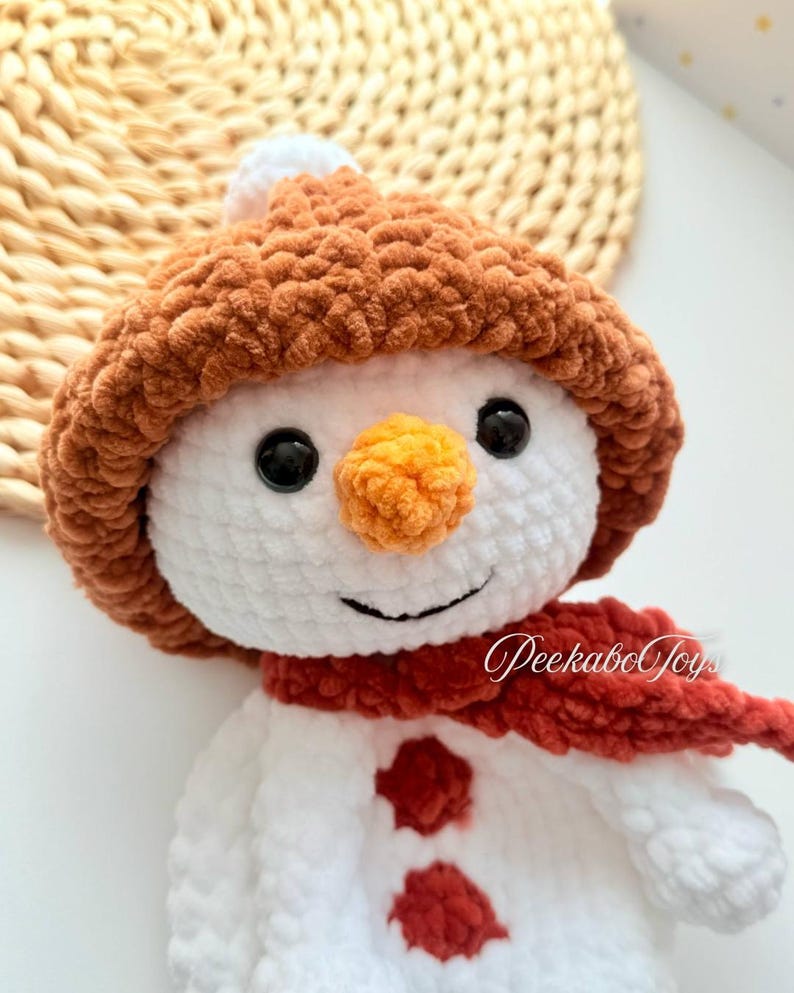 May include: A white crocheted snowman with a brown crocheted hat, a red crocheted scarf, and orange crocheted nose. The snowman has two red buttons.