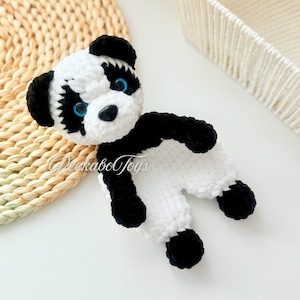 Puede incluir: Un pequeño juguete de panda de crochet con pelaje blanco y negro. El panda tiene ojos azules y una nariz negra. El juguete está sentado sobre una superficie blanca.