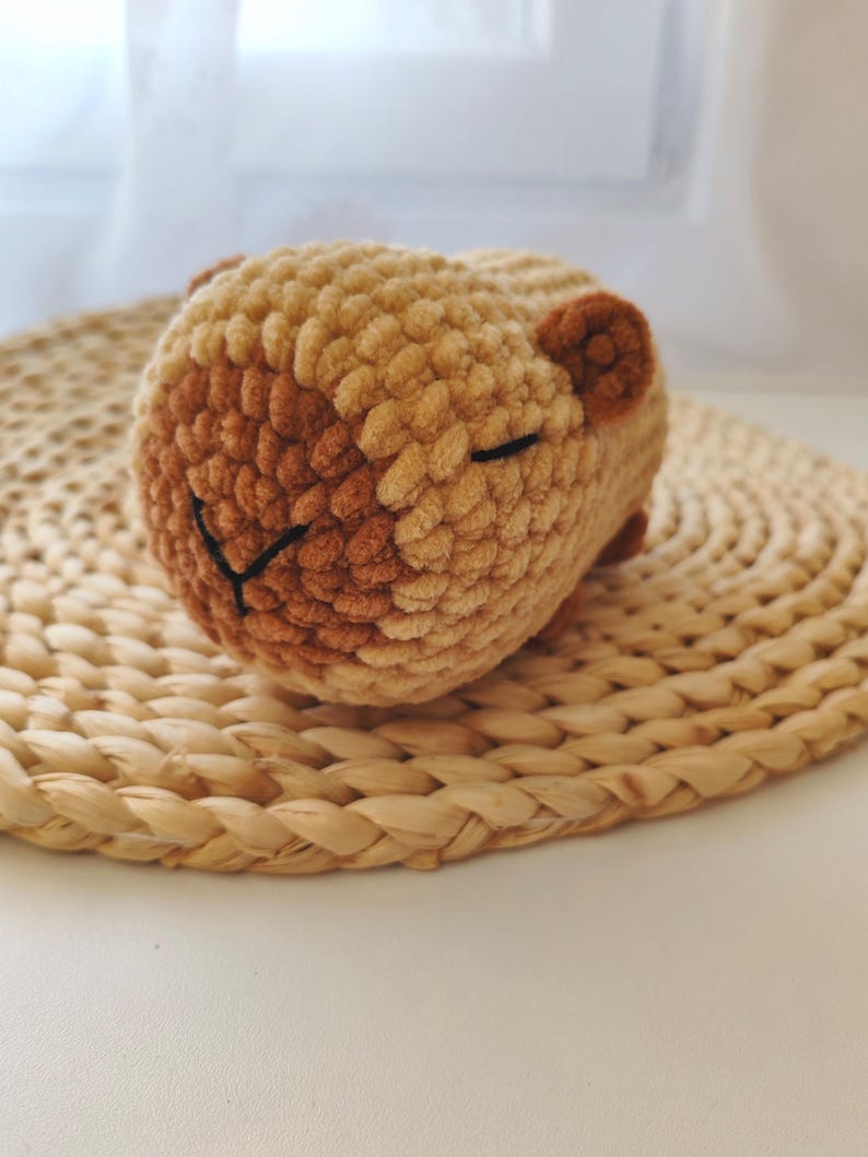 May include: A crocheted stuffed animal in the shape of a sleeping brown and tan guinea pig. The guinea pig is lying on a woven straw mat.