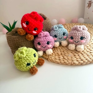 Puede incluir: Cinco peque&ntilde;as figuras de peluche tejidas a ganchillo, redondas y hechas de hilo suave y texturizado. Cada figura presenta ojos negros bordados, mejillas rosadas y una boca peque&ntilde;a. Los colores incluyen rojo, verde claro, morado, azul claro y rosa.