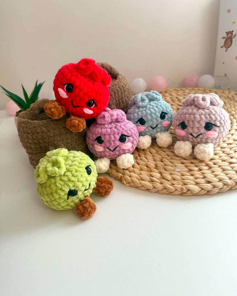 Puede incluir: Cinco peque&ntilde;as figuras de peluche tejidas a ganchillo, redondas y hechas de hilo suave y texturizado. Cada figura presenta ojos negros bordados, mejillas rosadas y una boca peque&ntilde;a. Los colores incluyen rojo, verde claro, morado, azul claro y rosa.
