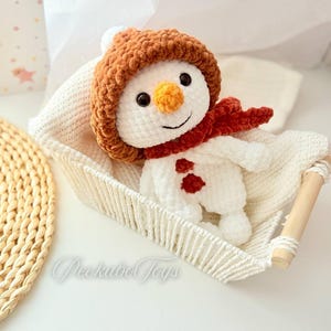 May include: A white crocheted snowman wearing a brown crocheted hat and a red scarf. The snowman is sitting in a white crocheted basket.