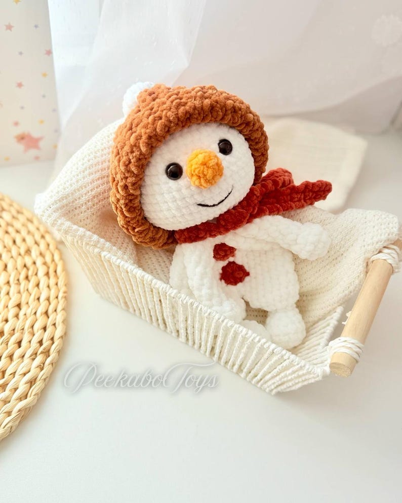 May include: A white crocheted snowman wearing a brown crocheted hat and a red scarf. The snowman is sitting in a white crocheted basket.