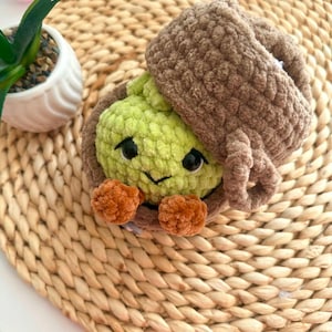 Puede incluir: Un juguete de peluche tejido a ganchillo con forma de criatura verde claro con ojos negros y una peque&ntilde;a sonrisa, metido dentro de una c&aacute;scara marr&oacute;n con textura en forma de bellota. La criatura tiene dos peque&ntilde;os pies naranjas redondeados.