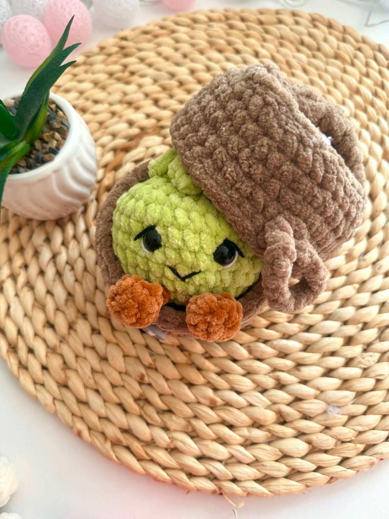 Puede incluir: Un juguete de peluche tejido a ganchillo con forma de criatura verde claro con ojos negros y una peque&ntilde;a sonrisa, metido dentro de una c&aacute;scara marr&oacute;n con textura en forma de bellota. La criatura tiene dos peque&ntilde;os pies naranjas redondeados.