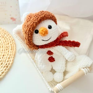 May include: A white crocheted snowman wearing a brown crocheted hat and a red scarf. The snowman is sitting in a white wicker basket.