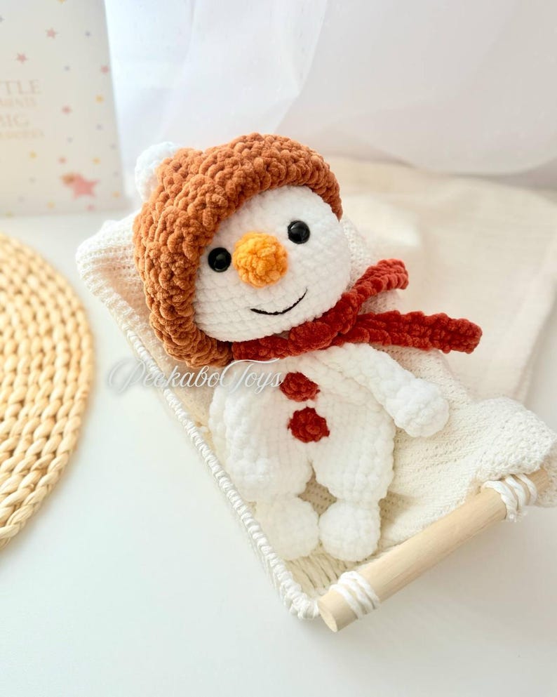 May include: A white crocheted snowman wearing a brown crocheted hat and a red scarf. The snowman is sitting in a white wicker basket.