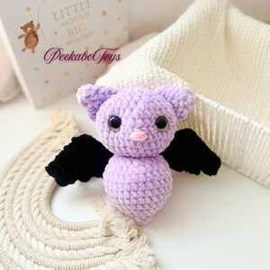 May include: A handmade lavender crochet bat with black wings and large, round, green eyes. The bat has a pink nose and small ears. The image also shows a decorative rainbow and a book with the text "Little Moments Big Memories" and the brand name "Peekabo Toys".