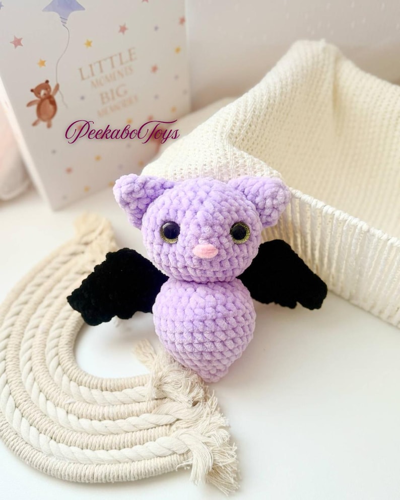 May include: A handmade lavender crochet bat with black wings and large, round, green eyes. The bat has a pink nose and small ears. The image also shows a decorative rainbow and a book with the text "Little Moments Big Memories" and the brand name "Peekabo Toys".