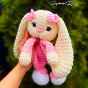 Può includere: Un giocattolo a forma di coniglietto all'uncinetto fatto a mano in crema, rosa e bianco. Il coniglietto ha lunghe orecchie flosce, un naso rosa e piccoli occhi neri. Le braccia e le gambe del coniglietto sono rosa e bianche. Il testo "PeekaboToys" è nell'angolo in alto a destra.
