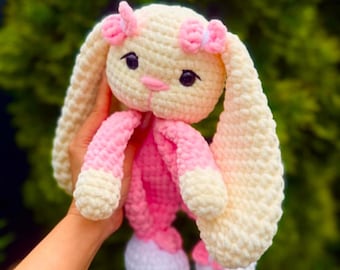 以下が含まれることがあります： クリーム、ピンク、白のハンドメイドかぎ針編みウサギのおもちゃ。ウサギは長く垂れた耳、ピンクの鼻、小さな黒い目を持っています。ウサギの腕と脚はピンクと白です。「PeekaboToys」の文字が右上隅にあります。
