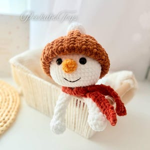 May include: A white crocheted snowman wearing a brown crocheted hat and a red crocheted scarf. The snowman is sitting in a white wicker basket.