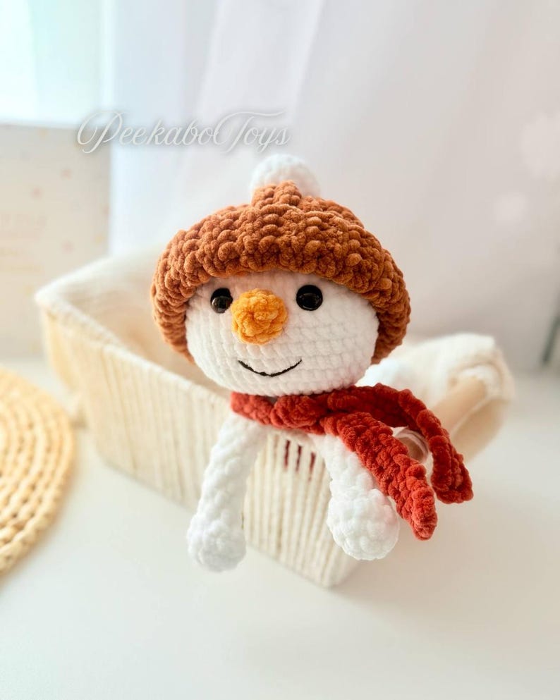 May include: A white crocheted snowman wearing a brown crocheted hat and a red crocheted scarf. The snowman is sitting in a white wicker basket.