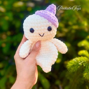 May include: A handmade, white crochet doll with a lavender hat and black eyes. The doll has a smiling face and small arms. The text "PeekaboToys" is visible in the upper right corner. The doll is held in a person's hand.