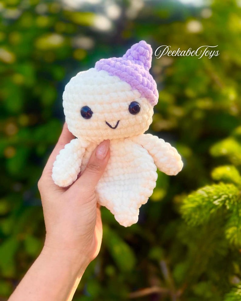 May include: A handmade, white crochet doll with a lavender hat and black eyes. The doll has a smiling face and small arms. The text "PeekaboToys" is visible in the upper right corner. The doll is held in a person's hand.