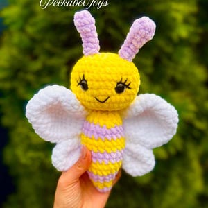 May include: A handmade crochet bee toy with yellow, lavender, and white details. The bee has large white wings, a smiling face, and the text "PeekaboToys" in cursive. The toy is held against a blurred green background.