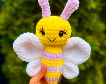 May include: A handmade crochet bee toy with yellow, lavender, and white details. The bee has large white wings, a smiling face, and the text "PeekaboToys" in cursive. The toy is held against a blurred green background.
