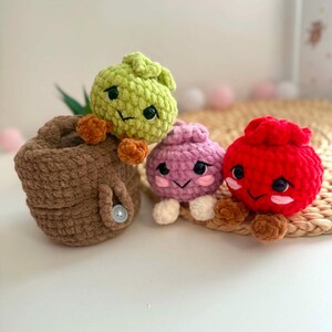 Puede incluir: Tres peque&ntilde;as figuras de ganchillo con caras bordadas y ojos de pl&aacute;stico negro. Una figura de color verde claro se sienta dentro de una cesta de ganchillo marr&oacute;n con un bot&oacute;n. Al lado hay una figura de color morado claro y una roja brillante.