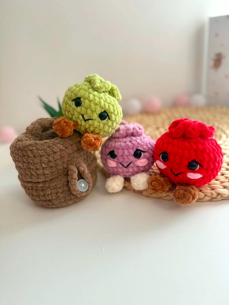 Puede incluir: Tres peque&ntilde;as figuras de ganchillo con caras bordadas y ojos de pl&aacute;stico negro. Una figura de color verde claro se sienta dentro de una cesta de ganchillo marr&oacute;n con un bot&oacute;n. Al lado hay una figura de color morado claro y una roja brillante.