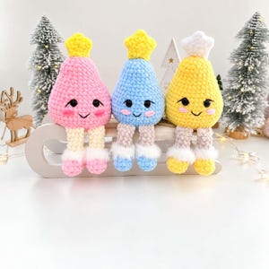May include: Three plush crochet dolls shaped like teardrops with smiling faces, rosy cheeks, and star-shaped toppers. The dolls are pink, blue, and yellow, each featuring dangling legs with white fuzzy cuffs, sitting on a white wooden sled.