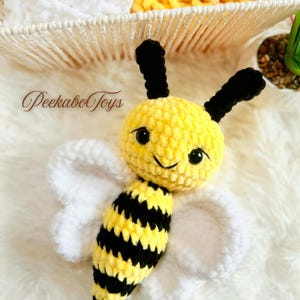 Può includere: Un giocattolo a forma di ape all'uncinetto giallo e nero fatto a mano con ali bianche. L'ape ha occhi e antenne neri. Il testo "Peekabo Toys" è nell'immagine. Il giocattolo è su una superficie bianca e soffice.