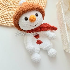 May include: A white crocheted snowman with a brown fuzzy hat and a red scarf. The snowman has black eyes, an orange nose, and a smiling mouth.