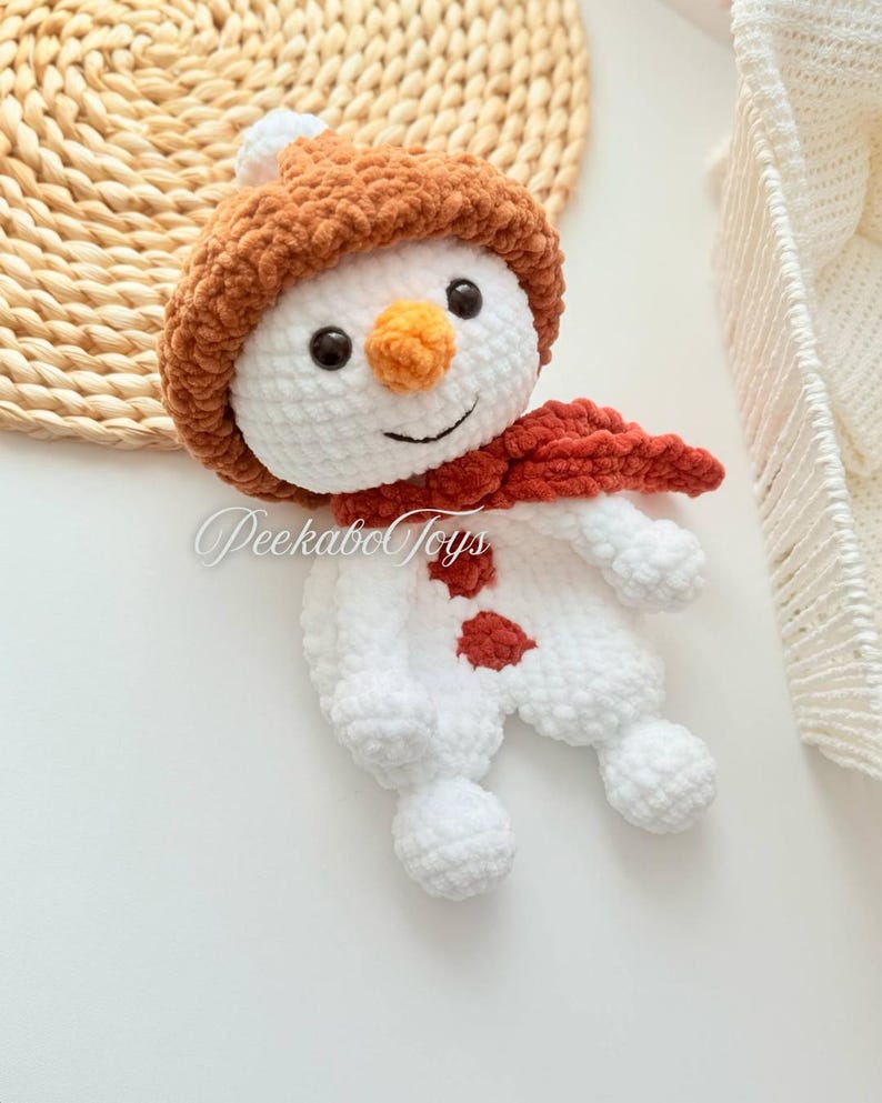 May include: A white crocheted snowman with a brown fuzzy hat and a red scarf. The snowman has black eyes, an orange nose, and a smiling mouth.