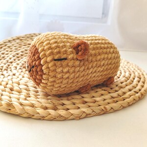 May include: A crocheted stuffed animal shaped like a sleeping bear. The bear is light brown with a darker brown nose and ears. It is lying on a woven straw mat.