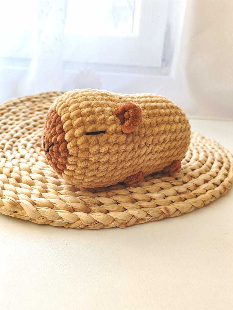 May include: A crocheted stuffed animal shaped like a sleeping bear. The bear is light brown with a darker brown nose and ears. It is lying on a woven straw mat.
