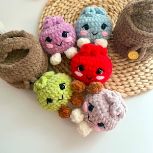 Puede incluir: Una colecci&oacute;n de cinco peque&ntilde;as criaturas de ganchillo de felpa, redondas, en colores morado, azul claro, rojo, verde lima y lavanda suave, cada una con ojos de seguridad negros, mejillas bordadas en rosa y extremidades peque&ntilde;as, junto a dos cestas de ganchillo marrones.
