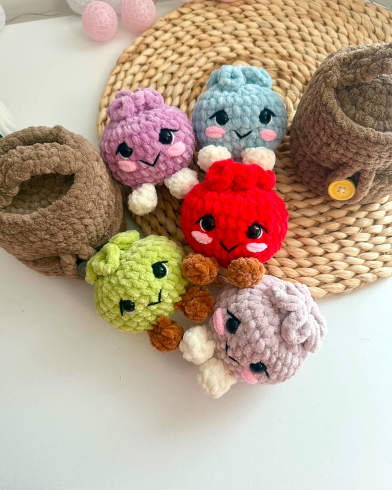 Puede incluir: Una colecci&oacute;n de cinco peque&ntilde;as criaturas de ganchillo de felpa, redondas, en colores morado, azul claro, rojo, verde lima y lavanda suave, cada una con ojos de seguridad negros, mejillas bordadas en rosa y extremidades peque&ntilde;as, junto a dos cestas de ganchillo marrones.