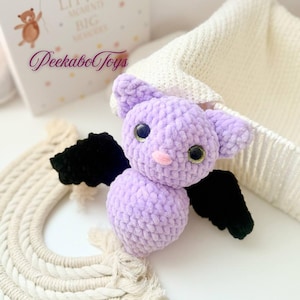 May include: A handmade, lavender crochet bat with black wings and large, glittery eyes. The plush toy has a pink nose and is part of a collection of PeekaboToys. A white knitted hat and a cream-colored macrame rainbow are in the background.