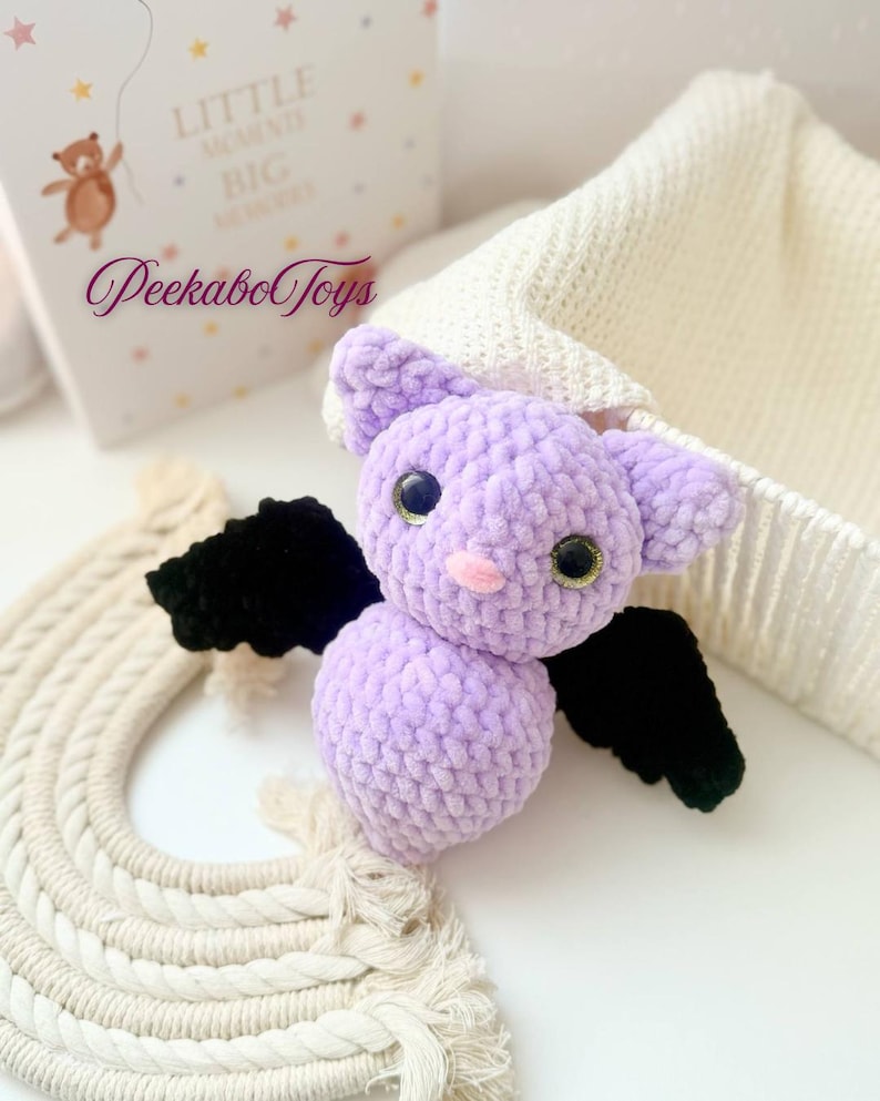 May include: A handmade, lavender crochet bat with black wings and large, glittery eyes. The plush toy has a pink nose and is part of a collection of PeekaboToys. A white knitted hat and a cream-colored macrame rainbow are in the background.