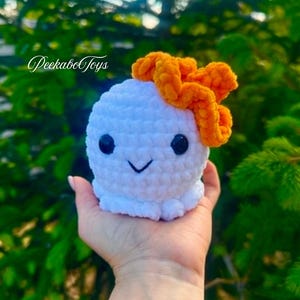 May include: A white crocheted octopus toy with black eyes and a smiling mouth, topped with an orange bow. The toy is held in a hand against a blurred green background. The text "PeekaboToys" is visible.