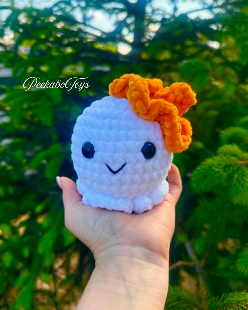 May include: A white crocheted octopus toy with black eyes and a smiling mouth, topped with an orange bow. The toy is held in a hand against a blurred green background. The text "PeekaboToys" is visible.