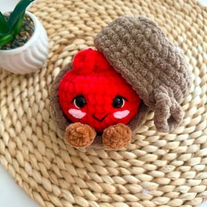 Puede incluir: Un juguete de peluche tejido a ganchillo que presenta un personaje redondo de color rojo brillante con ojos negros y mejillas rosadas, dentro de una c&aacute;scara marr&oacute;n texturizada con forma de bellota. Tiene pies peque&ntilde;os y el sombrero est&aacute; inclinado.
