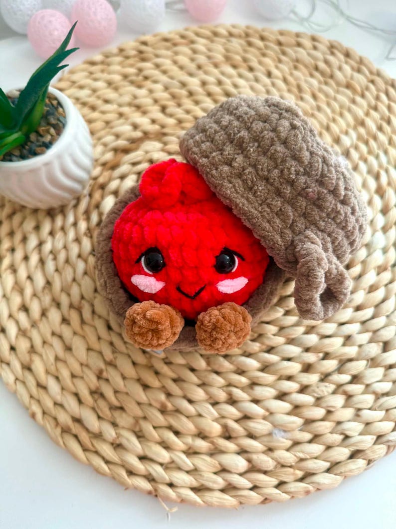Puede incluir: Un juguete de peluche tejido a ganchillo que presenta un personaje redondo de color rojo brillante con ojos negros y mejillas rosadas, dentro de una c&aacute;scara marr&oacute;n texturizada con forma de bellota. Tiene pies peque&ntilde;os y el sombrero est&aacute; inclinado.