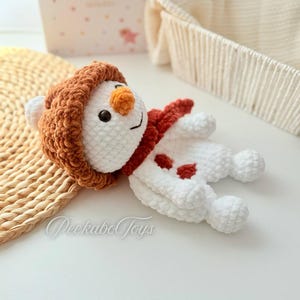 May include: A white crocheted snowman with a brown fuzzy hat and a red scarf. The snowman has a carrot nose and black eyes. The snowman is lying on a white surface.
