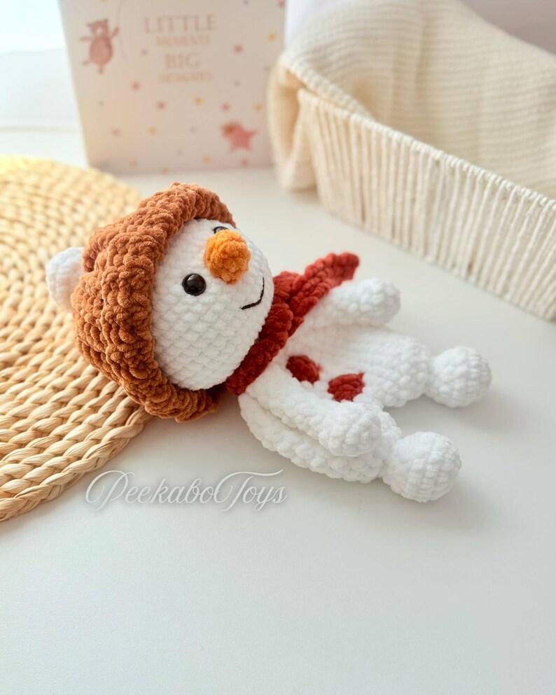 May include: A white crocheted snowman with a brown fuzzy hat and a red scarf. The snowman has a carrot nose and black eyes. The snowman is lying on a white surface.