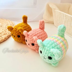 May include: Three handmade crochet cat toys in a row. The first is mustard yellow, the second is pink, and the third is mint green with pastel stripes. Each has black eyes and a small tail. The text "Peekaboo Toys" is in the lower left corner.