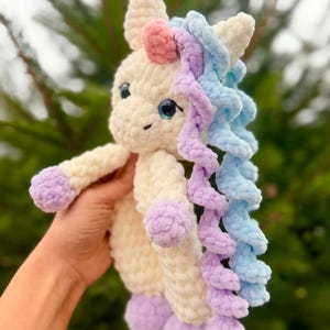 May include: A handmade, plush unicorn toy in cream, lavender, pink, and blue. The unicorn has a cream-colored body, lavender hooves, and a pink horn. Its mane is a mix of blue and lavender, and it has blue eyes.