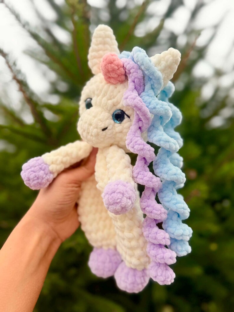 May include: A handmade, plush unicorn toy in cream, lavender, pink, and blue. The unicorn has a cream-colored body, lavender hooves, and a pink horn. Its mane is a mix of blue and lavender, and it has blue eyes.