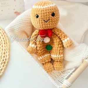 May include: A crocheted gingerbread man toy with a red bow, white icing details, and green and white buttons. The toy is lying in a white hammock.