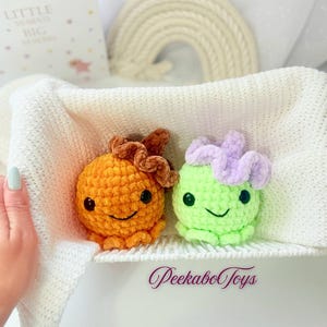 May include: Two handmade crochet octopus toys. One is orange with a brown flower, the other is green with a purple flower. Both have black eyes and smiling faces. The toys are in a white basket. The text "PeekaboToys" is visible.