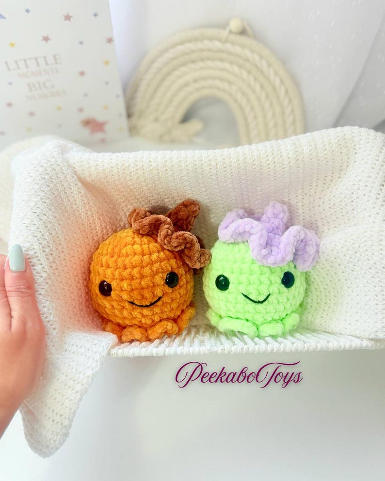 May include: Two handmade crochet octopus toys. One is orange with a brown flower, the other is green with a purple flower. Both have black eyes and smiling faces. The toys are in a white basket. The text "PeekaboToys" is visible.