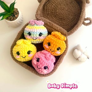May include: A brown crocheted basket containing four small, round, plush crocheted characters in yellow, orange, pink, and a pastel rainbow stripe. Each character features black plastic eyes, a stitched smile, and pink cheeks. Text reads Baby Dimple.